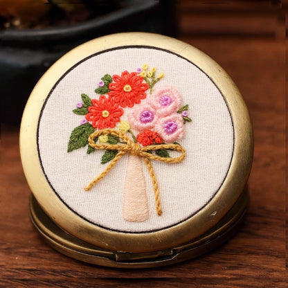 A charming hand-embroidered compact mirror featuring a delicate bouquet of coral-red and blush-pink flowers tied with a soft golden bow. Each stitch captures the warmth of spring mornings and the joy of blooming life. Elegant and petite, it’s a perfect companion for your handbag — a little bloom of happiness wherever you go.