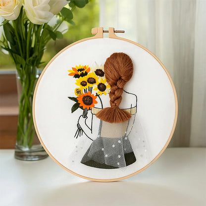 Embroidery hoop with a design of a girl with a braid holding sunflowers, placed on a surface with a blurred floral background.