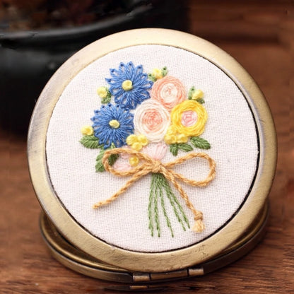 This lovely hand-embroidered compact mirror features a bouquet of blue daisies, yellow blossoms, and soft peach roses tied with a gentle golden bow. The design captures the freshness of spring mornings — vibrant yet soothing. Perfectly sized to slip into your purse, it brings a touch of handmade charm and floral serenity wherever you go.