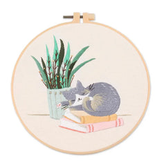 Embroidery hoop with a cat reading a book and a plant design on a white background